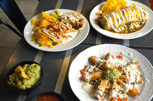 Three Plates Each on a Table, One with a Burrito and Chips, Another with Taquitos and Guac, and the Last with Three Tacos. A Bowl of Guac and Salsa Nearby