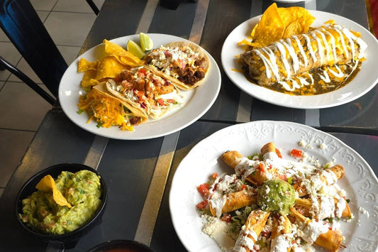 A burrito, a plate of tacos, and a plate of rolled tortillas next to a small bowl of guacamole