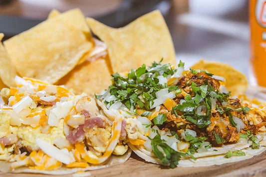 Two Tacos Topped with Cheese, Cilantro, and Onions on a Wooden Cutting Board with Tortilla Chips