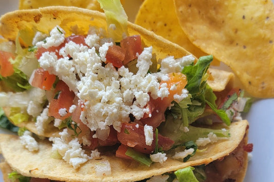 Close up of a hard shelled taco with lettuce, crumble cheese and tomatoes on a white plate with tortilla chips