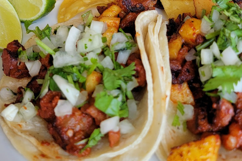 Best Mexican Food Delivery in Laguna Niguel close up of two tacos with meat, diced onion, and diced cilantro with lime wedges on a plate