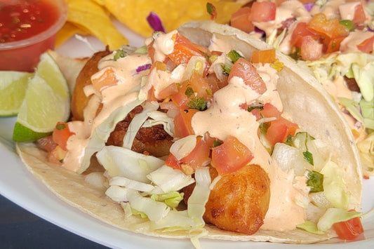 Close Up of Two Fish Tacos Filled with Fish Topped with Shredded Lettuce, Pico de Gallo, and Sauce with Two Lime Wedges and a Plastic Ramekin of Salsa