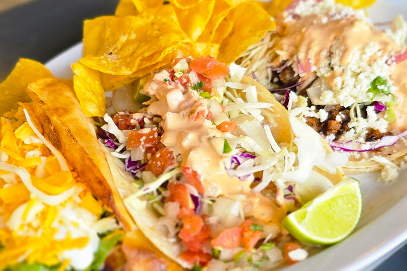 a Plate with Three Tacos Covered in Cheese, Shredded Lettuce, Lime Wedges and Tortilla Chips