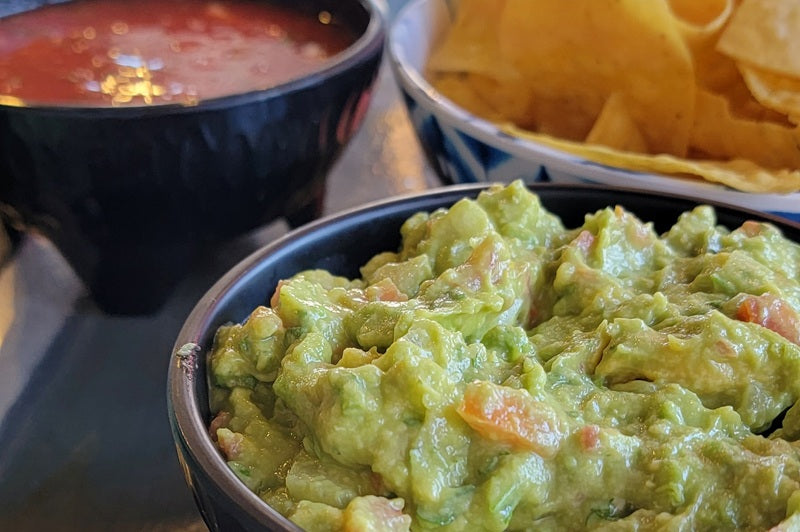 Close Up of a Small Bowl of Guacamole with a Small Bowl of Salsa and a Basket of Tortilla Chips in the Background