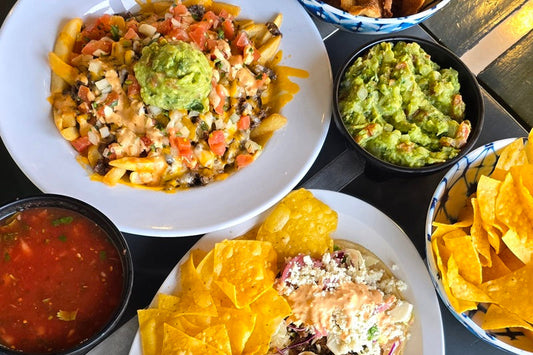 An Assortment of Plates on a Table One with Carne Asada Fries, One with Tacos and Rice and Beans, a Small Bowl of Guacamole, a Small Bowl of Tortilla Chips